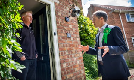 Adrian Ramsay, wearing a suit with a Green party rosette, speaks to a man who is standing in the doorway of a redbrick house