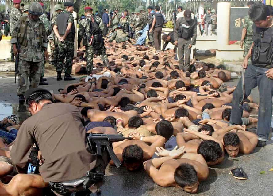 Dozens of shirtless men with their hands bound behind them lie face down surrounded by Thai police and soldiers