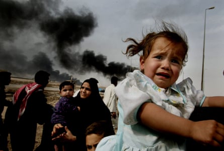 A crying preschool girl and a preschool boy are carried along a road with black smoke rising in the background
