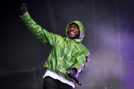 Skepta dressed in a bright green wet-look hooded Stone Island jackets miles and lifts his gloved hand in the air while performing on stage