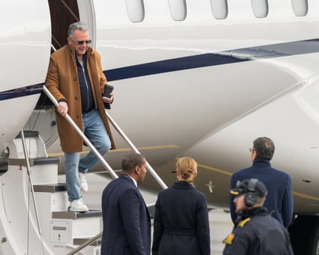 Steve Witkoff descends aircraft stairs holding a coffee cup, greeted by waiting officials