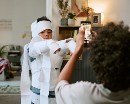 A child wrapped in toilet paper raises their arms while a sibling photographs them on a phone