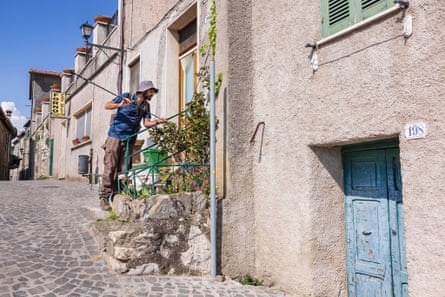 A man stands next to a house on a sunny cobbled street holding a fishing rod