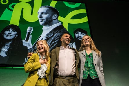 Spencer, Polanski and Berry stand on stage in front of a big banner with pictures of Polanski and other party figures. They are smiling and looking jubilant.