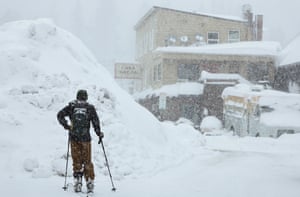 A person pauses on their skis beside a large pile of snow, looking towards a building as snow falls