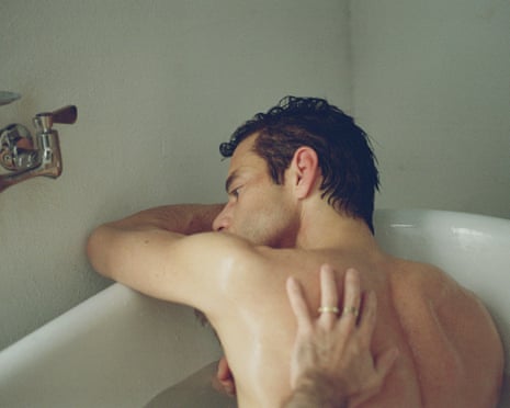 A shirtless man leans against a bathtub edge while a hand wearing a ring touches his back