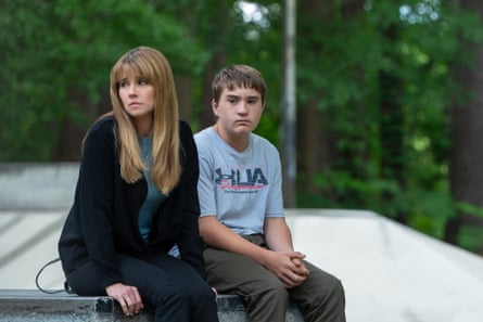 A woman with long blond hair and a fringe sits with a teenage boy both looking glum with trees in background