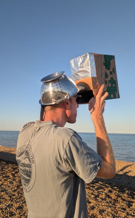 Matt stands on a beach with a colander on his head, looking into a box with tinfoil taped to it