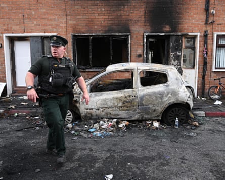 A Northern Ireland police officer stands in front of a torched car and burned houses.