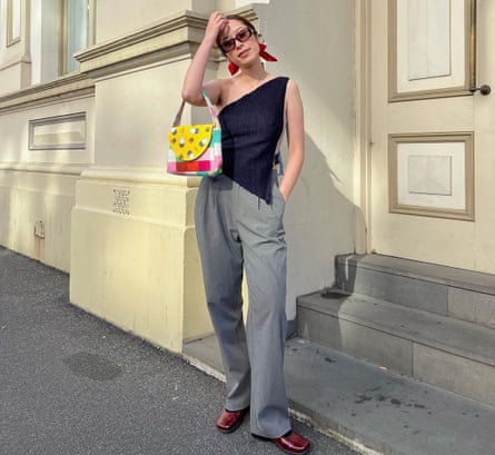 Woman wearing a black top and grey pants with a colourful bag on her arm stands outside a cream building