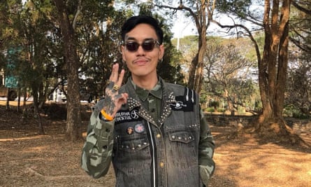 A man wearing a khaki shirt, grey denim vest and sunglasses holds up his index, middle and ring fingers.