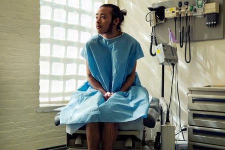 A genderqueer person sitting in a hospital gown sitting in an exam room.