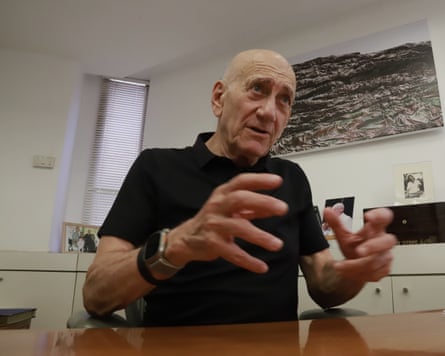 he sits behind a long wooden table or desk with white cupboards and a painting behind him. He is nearly 80, bald, and wears a black polo shirt; he is raising his hands as he speaks.