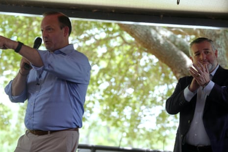 man in blue button-down speaks into microphone as another man claps behind him