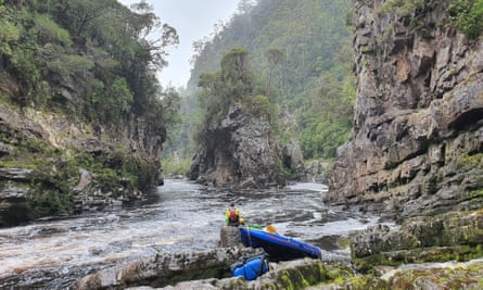 Oliver Cassidy is sitting on a rock on the bank of the Franklin River, looking out over the rock island bend. His raft is behind him on the bank