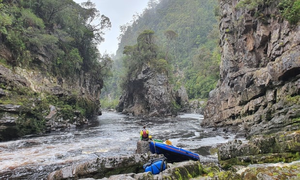 Oliver Cassidy is sitting on a rock on the bank of the Franklin River, looking out over the rock island bend. His raft is behind him on the bank