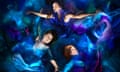 Three women lie with arms outstretched and eyes closed in floaty blue and indigo dresses