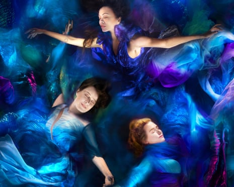Three women lie with arms outstretched and eyes closed in floaty blue and indigo dresses