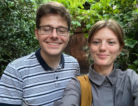 A young man with glasses and short brown hair, wearing a striped polo, and a young woman with dark blond hair tied back, wearing a grey shirt, standing by a fence with shrubs in front of it
