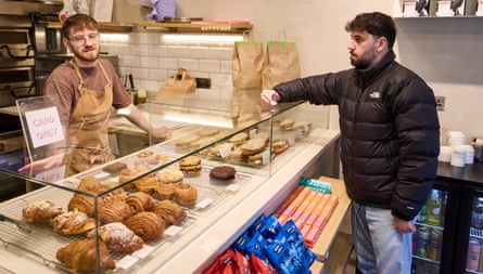 Sammy Gecsoyler trying to pay a barista for a coffee at a pastry counter in a cafe –-with a Card Only sign visible to the side