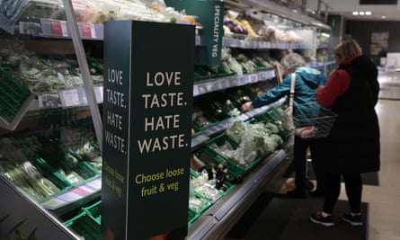 A shopper in a Waitrose fresh produce section with a sign that reads 'Love taste, hate waste: choose loose fruit and veg'