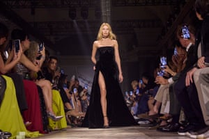A woman in a strapless long black dress walks down the runway as people on either side take pictures with their mobile phones