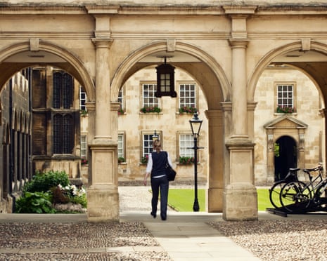 A person in dark clothing walks through a Cambridge University stone archway toward a courtyard.