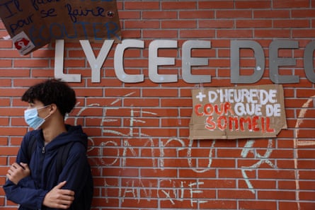 Student wearing medical face mask, leans against the graffiti-strewn brick wall of the school, on which the sign (in French) has been attached