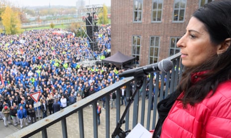 Woman wears red jacket and speaks into microphone on balcony with thousands of people below here