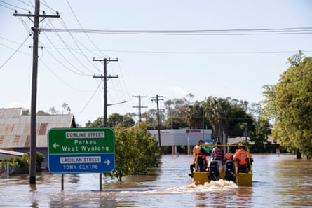 A small yellow rescue move travels through flood waters in a street under powerlines. People in the boat are wearing orange hi-vis and there is a road sign to Parkes and West Wyalong and the town centre