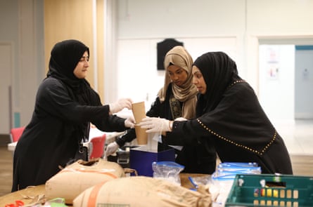Three women wearing hijabs stand at a table measuring out sugar to put into bags.