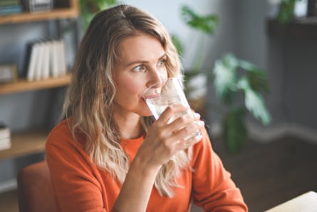 A blond woman wearing orange top drinks a glass of water while sitting down.