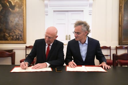 Alex Karp and John Healey sign open pages of documents as they sit at a long table in a Whitehall office.