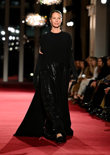 Christy Turlington, dressed in elegant black outfit, walks a red runway.