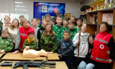 A screenshot of a deleted image showing Russian Red Cross staff posing with Kalashnikov rifles at a military event for children. 