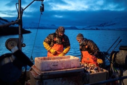Two men in protective clothing, hats and gloves on the deck of a boat in half-light