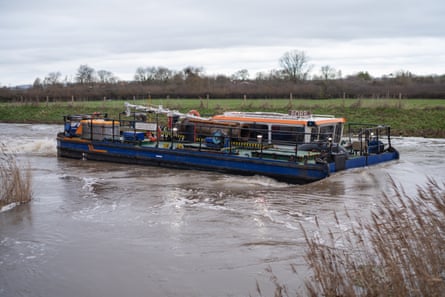 A low, dark blue dredger with flat deck moves along the brownish river; the water level is high. There are flat green fields in the background, grasses and reeds in the foreground, and the sky is grey.