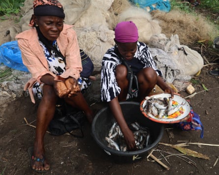 ‘The water is no longer our friend’: how dredging is pushing Lagos Lagoon towards ecosystem collapse – photo essay Two women sit on the ground next to a large bowl of fish. One holds a plate of fresh fish in her hand.
