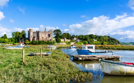 A landscape photograph of Laughlarne Castle, which is seen in the distance. The sky is blue, the trees are a vivid green. In the foreground are boats on the River Tar.