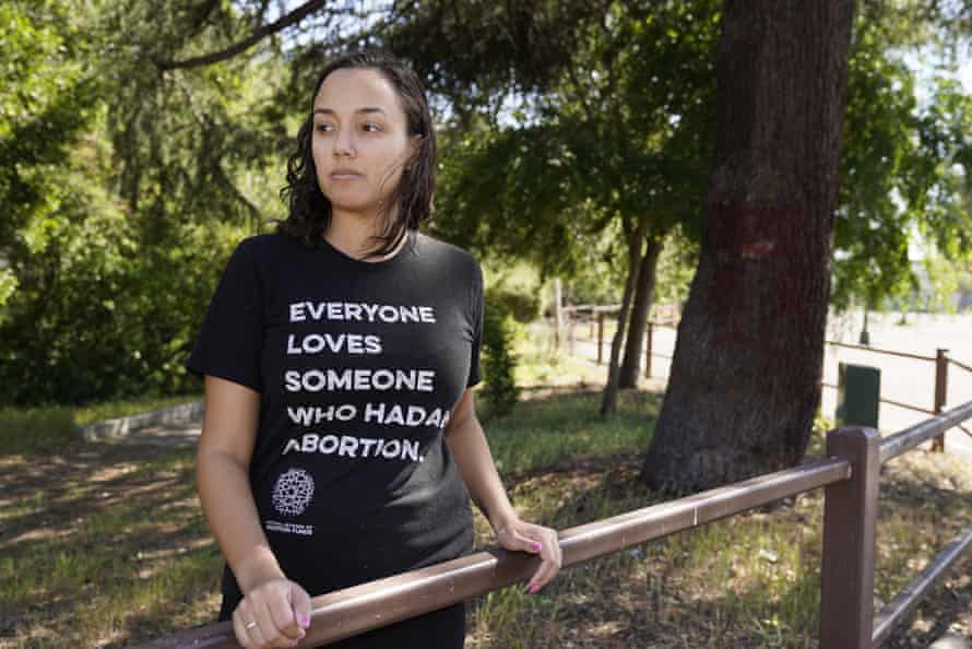 A woman stands next to a fence in a park wearing a shirt that says 'Everyon loves someone who had an abortion'.