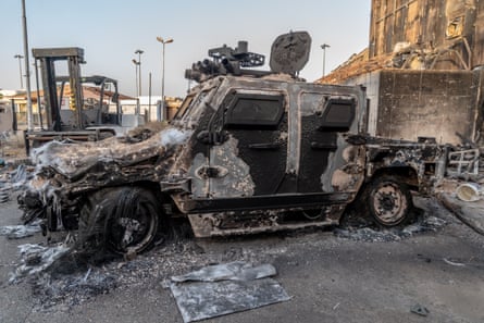 A burned military vehicle on the tarmac outside a building.