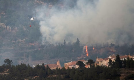 A helicopter flies over a village near a wooded area that is on fire