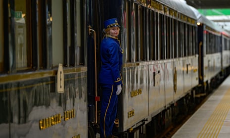 A young woman, probably a guard or an attendant, in a blue railway uniform, standing at the door of a vintage train carriage standing on a platform , looking back towards the camera