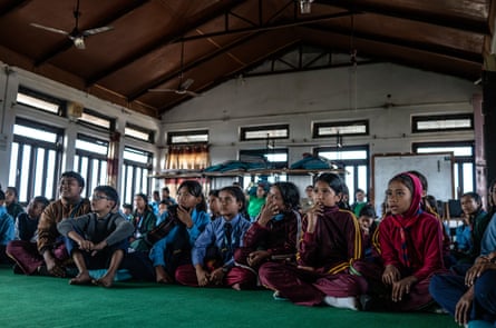 A large group of children sit on the floor of a large building