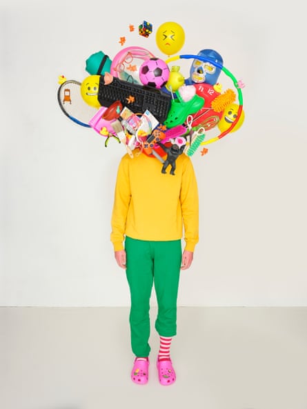 Photomontage of a body in a yellow sweatshirt and green sweatpants, one leg pulled up showing a pink and white stripy sock, pink crocs, and with a mass of objects – balloons, a keyboard, toilet plunger, tennis racket etc – in place of a head
