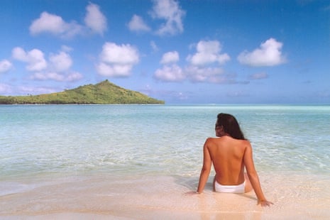 Woman in white bikini bottoms, back to camera, sitting on a sandy beach looking out to sea, blue sky and an island ahead of her
