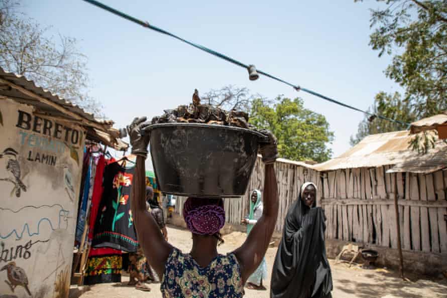 A woman walks with the tub of oysters balanced on her head along an unpaved road with huts on either side