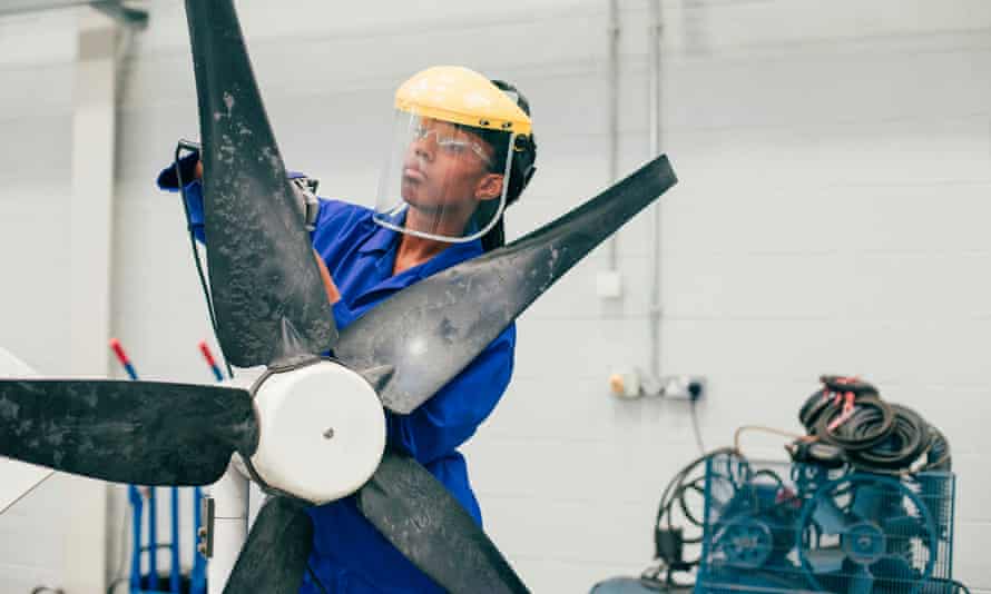 A young woman is working on a wind turbine in an engineering workshop. She is wearing a protective mask as well as blue coveralls.