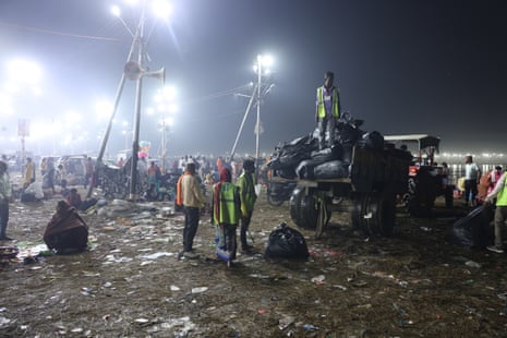 A man in hi-vis stands on the back of a truck covered in full black bags. Others also in hi-vis stand next to the truck. Other people and floodlights can be seen behind