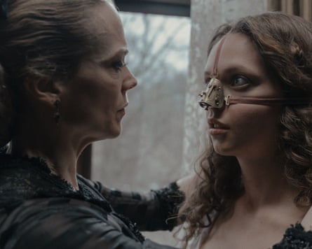 A woman purses her lips as she puts her arms on either side of a young woman with curly hair who looks terrified strapped into a curved device on her nose